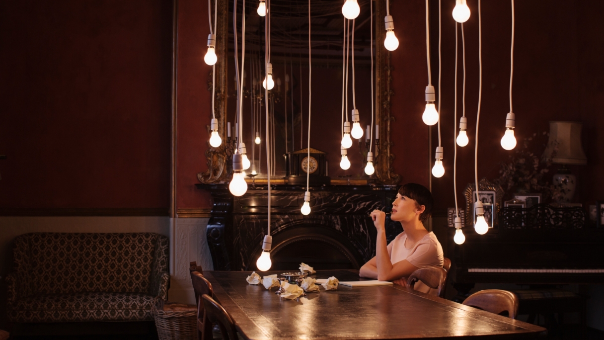 Woman chewing a pencil under a chandelier of light bulbs